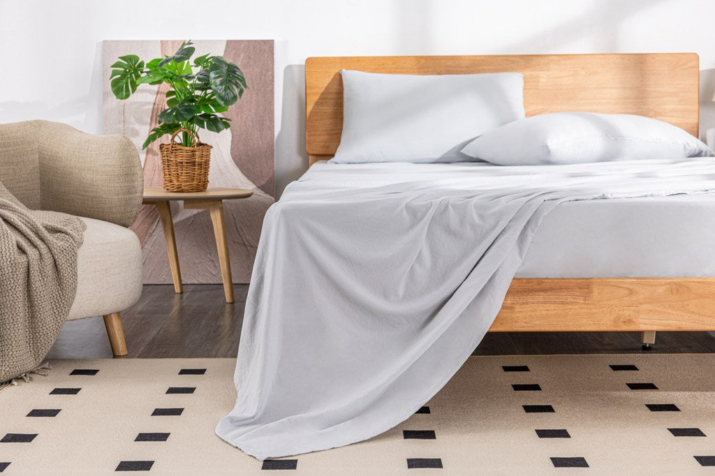 Bedroom with a bed covered in white Mlily bedding, wooden headboard, and plant on a side table.